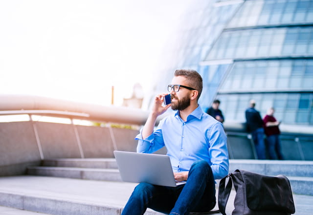 Mann im Business-Outfit sitzt mit Laptop auf dem Knie einer Stufe und telefoniert mit dem Handy