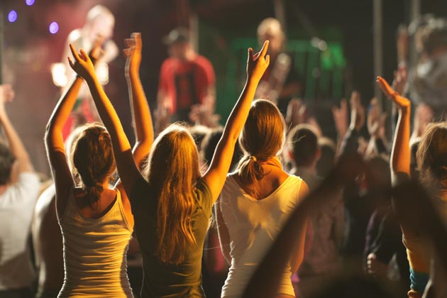 Rückansicht eines Publikums, das mit erhobenen Händen bei einem Open-Air-Musikfestival im Dunkeln vor einer Bühne steht