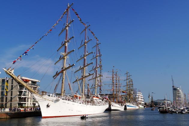 Mehrere weiß lackierte Segelschiffe mit eingezogenen Segeln liegen unter blauem Himmel in Bremerhaven vor Anker.