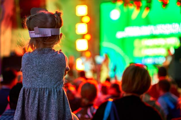 Rückansicht einer Menschenmenge vor einer Bühne bei einem Konzert, im Fokus ein kleines Mädchen auf den Schultern einer Person