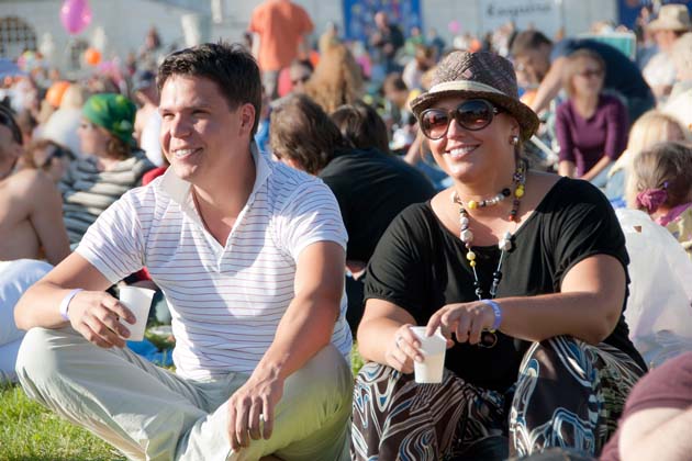 Zwei Personen sitzend fröhlich auf einer Wiese bei einem Festival, um sie herum weitere Personen