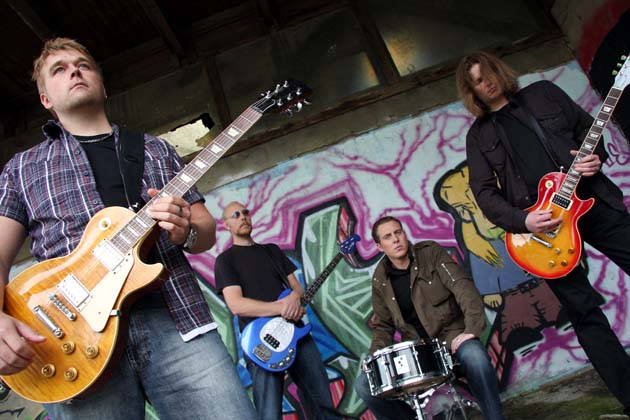 Band, bestehend aus 3 Gitarristen und einem Schlagzeuger, posiert vor einer Grafitti-Wand.