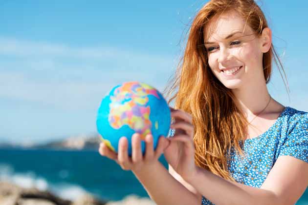 Eine junge Frau mit orangenen Haaren und blauer Bluse hält am Meer einen Globus in der Hand.