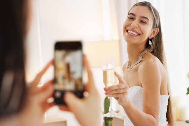 Eine Frau macht ein Foto mit dem Smartphone von ihrer Freundin im Brautkleid mit Sektglas in der Hand.