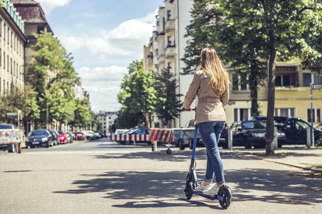 Von hinten fotografierte junge Frau, die mit beiger Jacke auf einem E-Scooter auf einer Straße umsäumt von Häusern in einem Wohngebiet fährt