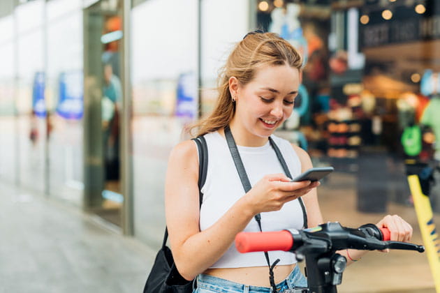 Junge blondhaarige Frau scannt mit ihrem Smartphone in der Hand den QR-Code eines E-Rollers, um ihn zu entsperren