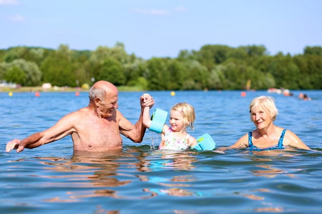 Glückliches, kleines Mädchen mit blauen Schwimmflügeln lernt im See gemeinsam mit ihren Großeltern schwimmen und hält dabei die Hand ihres Opas  
