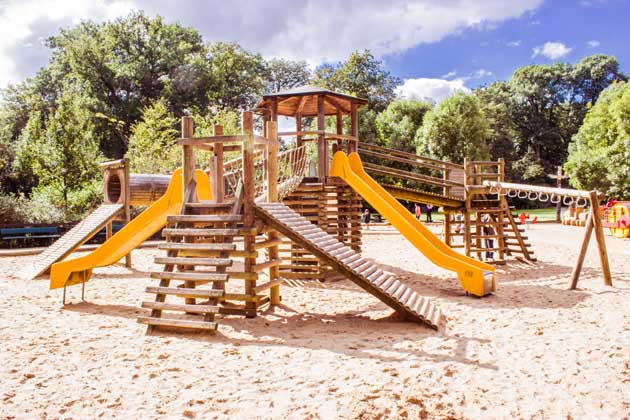 Spielplatz mit hölzernem Klettergerüst und gelben Rutschen auf Sandboden in einem Park.