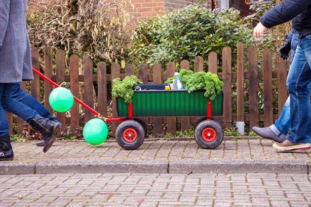 Menschen auf einer Kohltour ziehen einen grünen Bollerwagen mit Ballons an einem braunen Zaun vorbei.