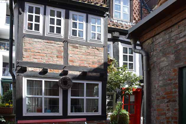 Fachwerkhausfassade aus roten Backsteinen und braunen Holzstämmen mit kleinen weißen Fenstern, einer knallroten Tür und einer roten Bank davor.