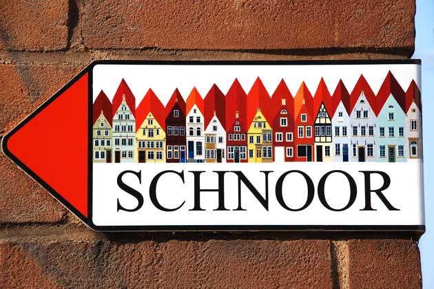 Rote Backsteinmauer an der ein pfeilförmiges Schild mit dem Titel "Schnoor" und einer bunten engstehenden Häuserreihe als Motiv drauf hängt.