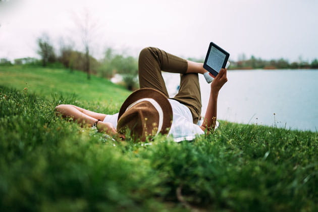 Eine Person liegt bequem im grünen Gras am Ufer des Werdersees, trägt einen Hut, hat die Bein überschlagen und liest in einem E-Book-Reader