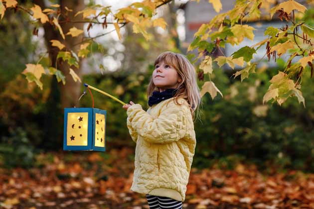 Ein kleines Kind steht mit einer Laterne unter einem Laubbaum im Herbst.