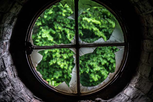 Ein kaputtes Kreisfenster mit Scherbenresten im Rahmen und Blick auf einen Busch.