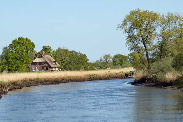 Eine Flusskurve der Wümme mit Gras, Bäumen und einem Haus mit Reetdach im Bremer Blockland.