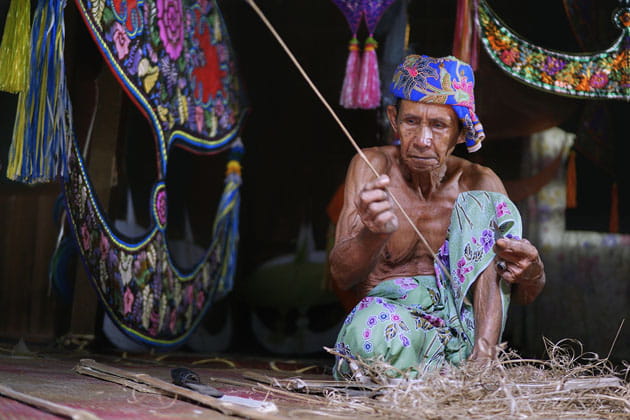Älterer Mann arbeitet in traditioneller Kleidung auf dem Boden einer Hütte umgeben von bunten Stoffen