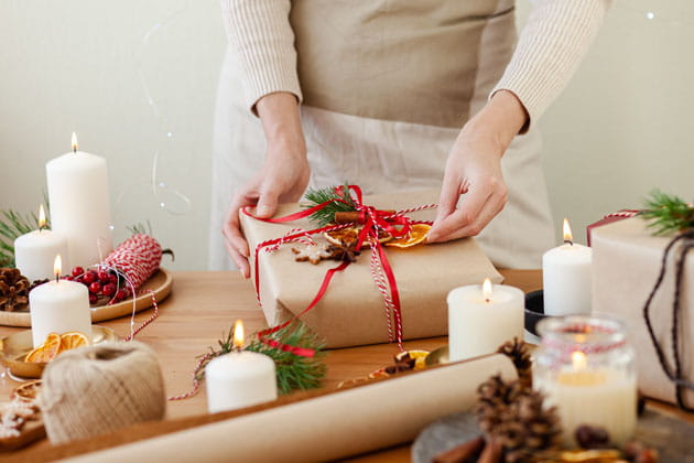 Junge Frau verpackt nachhaltige Weihnachtsgeschenke mit recycelbarem Geschenkpapier bei Kerzenschein und dekoriert sie mit getrockneten Orangen, Tannenzweigen und Bändern