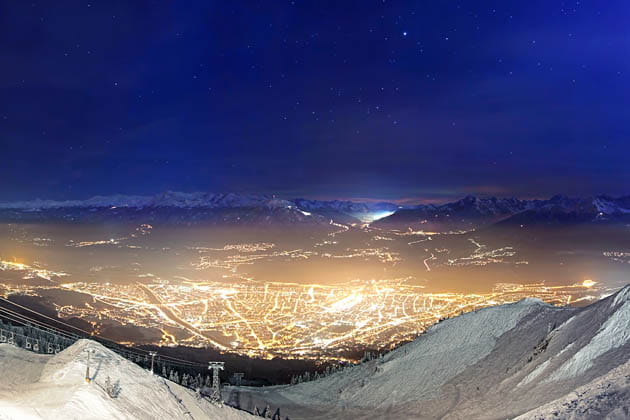 Lichtverschmutzte Stadt im Tal bei Nacht mit schneebedeckten Bergen im Vordergrund.