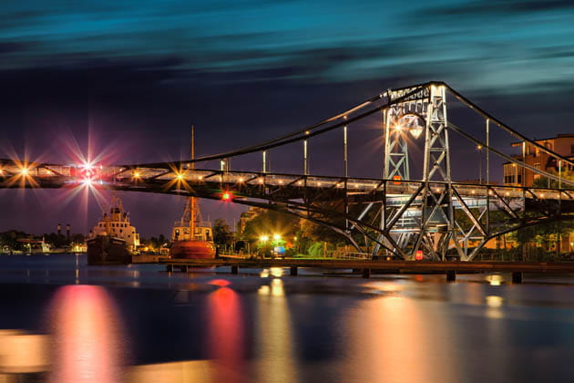 Eine hell beleuchtete Brücke über einem Fluss mit Schiffen im Hintergrund.