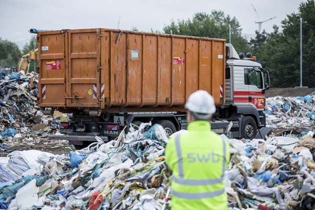 LKW, der Müll zu einer Mülldeponie liefert, während ein swb-Mitarbeiter in Rückenansicht dies beaufsichtigt