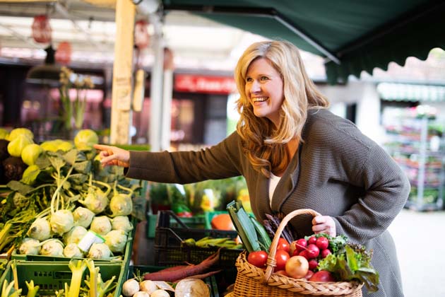Blondhaarige Frau mittleren Alters kauft frisches Gemüse auf einem lokalen Markt und sammelt die Einkäufe in einem Korb