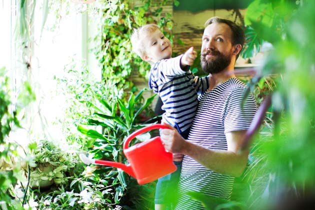 Mann mit Vollbart gießt mit einer roten Gießkanne die Pflanzen zu Hause, während er einen kleinen Jungen auf dem Arm trägt, der fasziniert auf etwas deutet