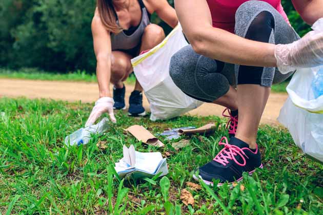 Zwei junge Frauen in Sportkleidung betreiben Plogging, d.h. sie sammeln Müll vom Wanderweg auf.