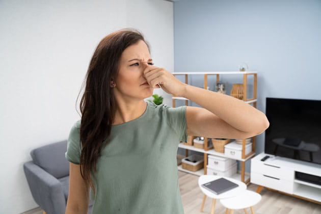 Braunhaarige, junge Frau in einem grünen T-shirt hält sich aufgrund eines schlechten Geruchs in ihrem Wohnzimmer ihre Nase zu. 