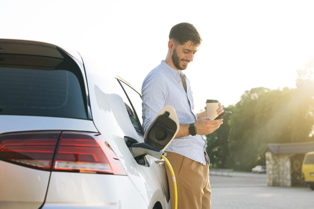 Mann mit Kaffee und Smartphone in der Hand lehnt sich an sein ladendes Plug-In-Hybrid-Auto.