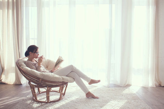 Frau mit Dutt und Brille entspannt in einem gemütlichen Sessel mit überschlagenen Beinen in ihrem Heim, während sie einen Tee trinkt und ein Buch liest