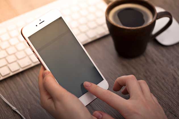Ein Finger bedient ein iPhone, während eine Tasse mit Kaffee, eine Tastatur und eine Maus im Hintergrund liegen.