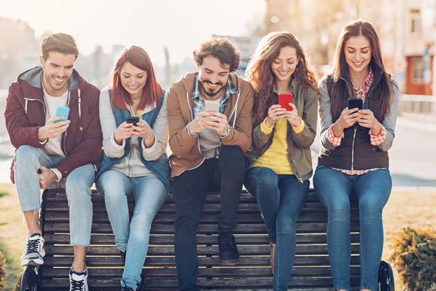 Eine Gruppe von jungen Menschen sitzt auf einer Parkbank und gucken fröhlich auf ihr Smartphone.