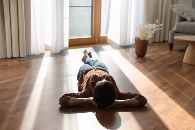 Eine junge Frau liegt mit dem Rücken auf dem beheizten Boden ihres Wohnzimmers, während durch die Vorhänge das Sonnenlicht scheint.