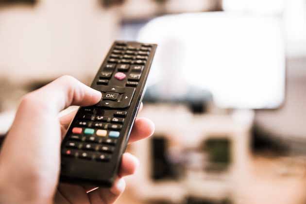 One hand holds the remote control, pointed at the television in the background.