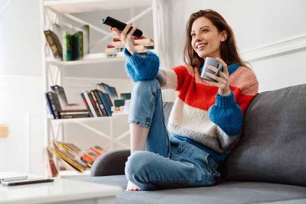 A young woman sits on a couch with a cup and remote control in her hand, standing in front of a white bookshelf.