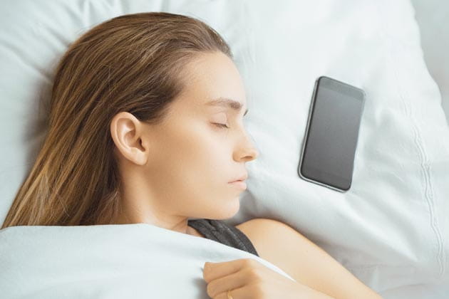Eine Frau liegt im Bett mit ihrem Smartphone neben ihrem Kopf auf dem Kopfkissen.