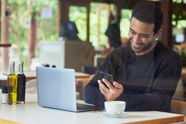 Ein junger Mann sitzt in einem Café und surft via WLAN mit Smartphone und Laptop im Internet.
