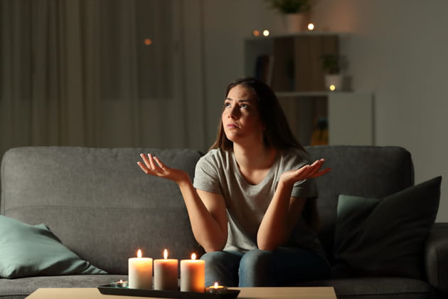 Eine junge Frau sitzt während eines Stromausfalls bei Kerzenschein in ihrem Wohnzimmer und macht eine verwunderte Geste.