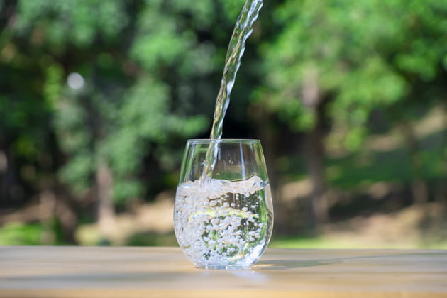 Ein Wasserglas steht draußen auf einem Tisch und wird mit Trinkwasser vor grüner Kulisse befüllt