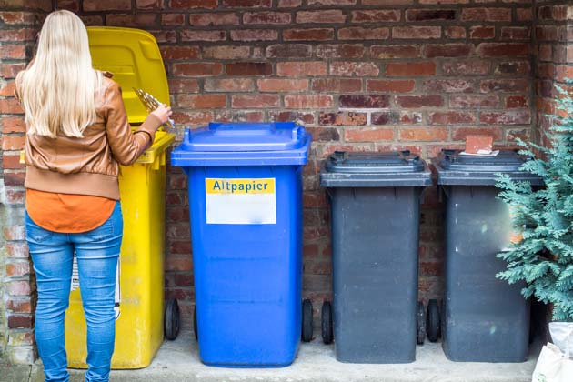 Eine blonde Frau trennt ihren Müll, indem sie ihren Abfall in die Gelbe Tonne wirft, neben dereine Altpapiertonne und zwei Restmülltonnen stehen