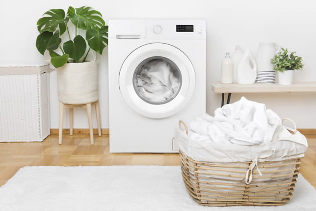 Abbildung einer weißen Waschmaschine, in der sich eine Ladung Wäsche befindet und ein voller Wäschekorb davor