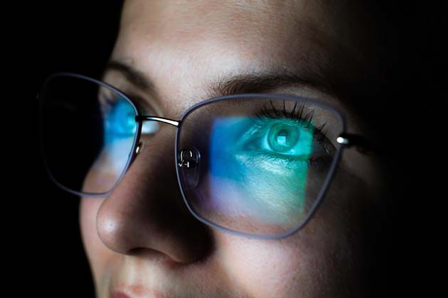 Nahaufnahme einer jungen Frau, die schräg nach vorne blickt und eine Blaulichtfilter-Brille trägt, in der sich ein Bildschirm spiegelt