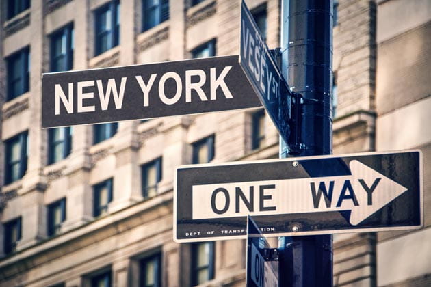Ein schwarzer Laternenmast mit den Schildern "New York" und "One-Way" dran vor einer Häuserfassade.