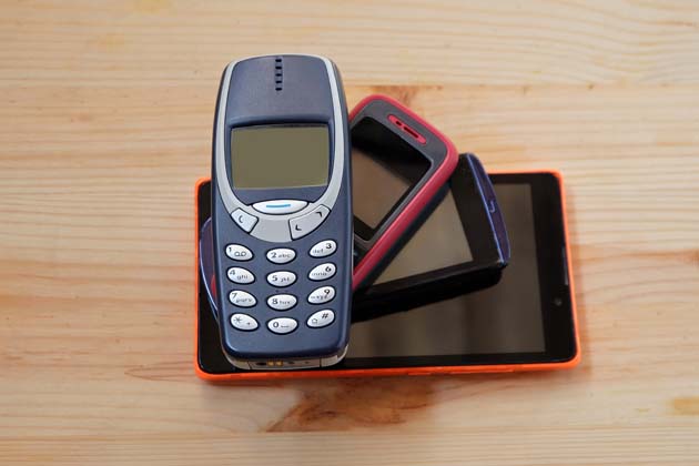 Ein Stapel aus 4 Handys auf einem Holztisch, ganz oben liegt ein altes Nokia-Handy und ganz unten ein Smartphone.