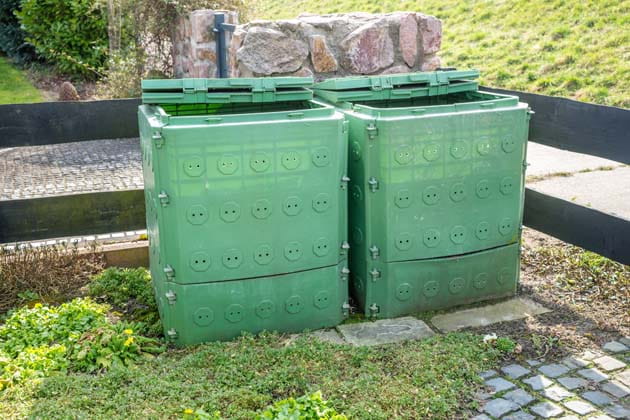 Zwei grüne Kompostbehälter nebeneinander vor einem kleinen Zaun; dahinter ein Weg und ein Deich