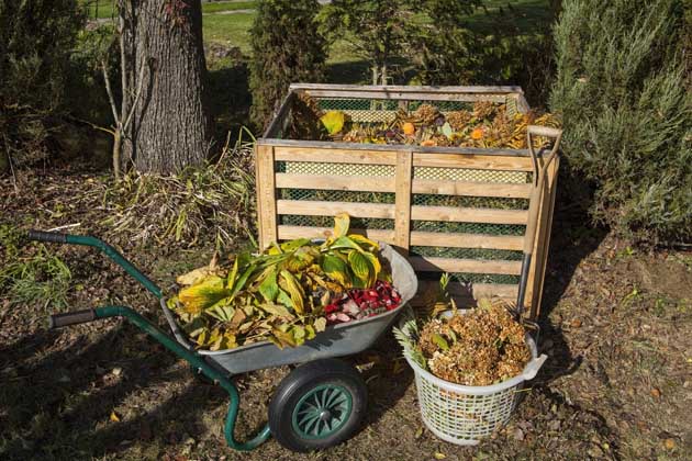 Mit Gartenabfällen gefüllte Schubkarre und Korb mit organischen Abfällen, die vor einem Kompost aus Holz im Garten stehen