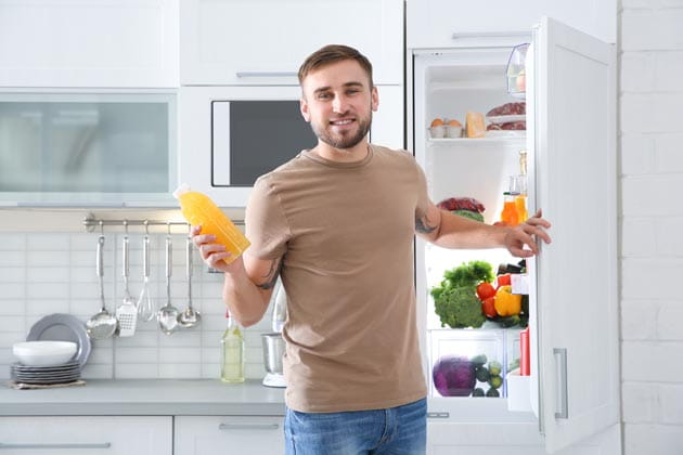 Ein Mann steht vor dem halbgeöffneten Kühlschrank in der Küche und wendet sich zum gehen mit einer Flasche O-Saft in der Hand.