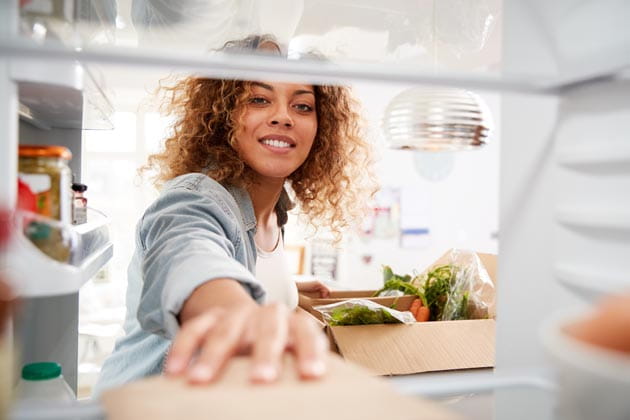 Blick aus dem Kühlschrank heraus auf eine Frau mit gelockten Haaren, die dabei ist ihre Lebensmittel in den Kühlschrank einzuräumen.