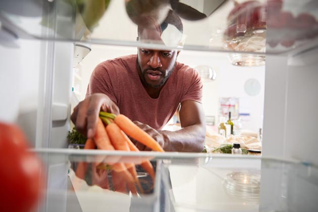 Blick aus dem Kühlschrank auf einen Mann, der gerade dabei ist, einen Bund Karotten in den Kühlschrank einzuräumen.