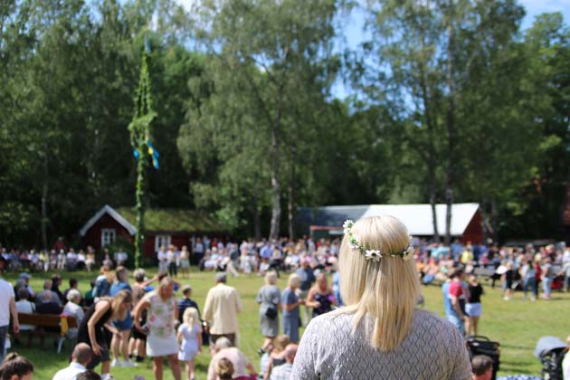 Rückenansicht einer jungen blondhaarigen Frau mit Blumenkranz im Haar auf einem traditionellen schwedischen Mittsommerfest mit einem Maibaum und feiernden Menschen im Hintergrund